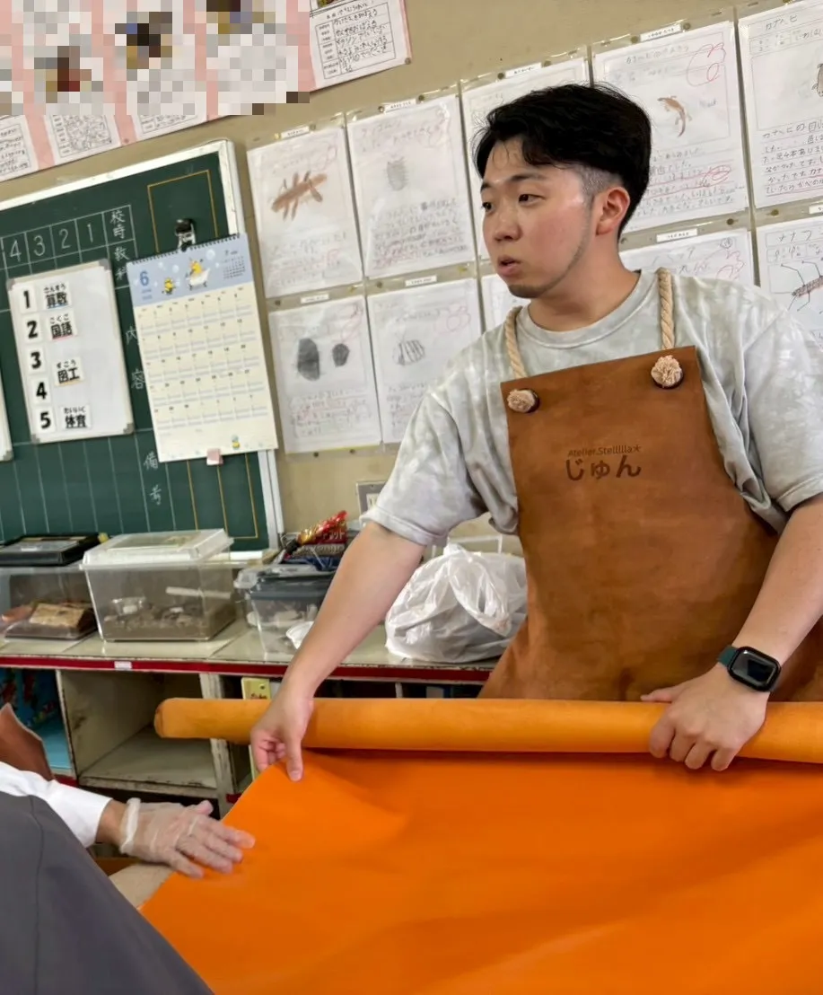 学校の教室で、Atelier Stelllllaの作り手が大きなオレンジ色の革を広げ、参加者が手で革に触れている様子